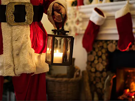 Â Santa Claus holding a lantern with stockings hanging above a fireplace in the background