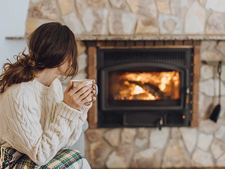 A woman is drinking coffee beside an indoor wood fireplace.